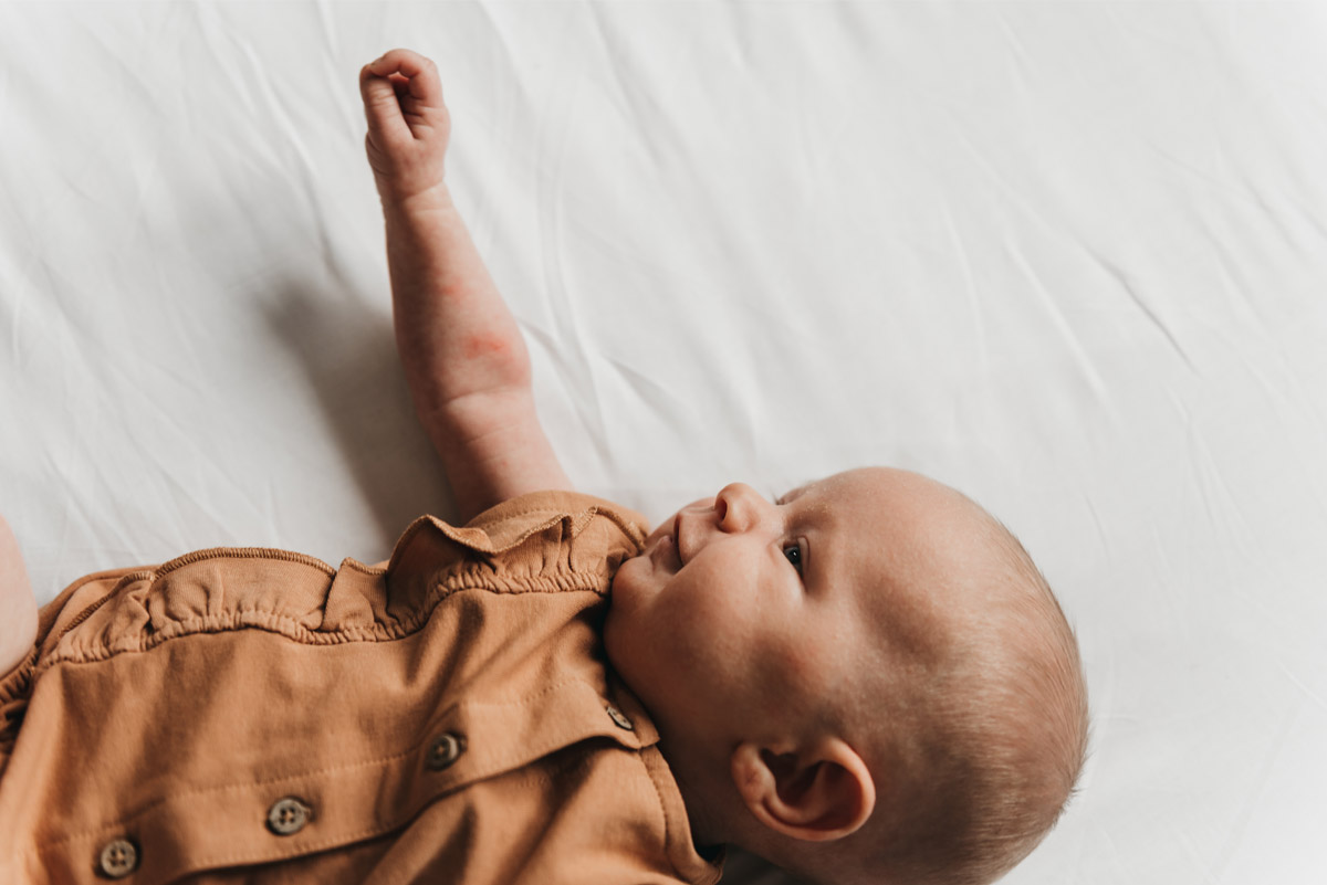 Baby in een bruin pakje ligt op bed en strekt zijn armpje omhoog